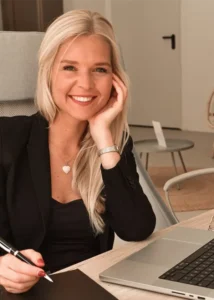 Portrait: Caroline Birke mit langen blonden Haaren sitzt lächelnd an einem Schreibtisch mit Notebook und Laptop und hält einen Stift in der Hand.