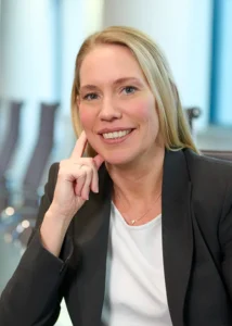 Eine lächelnde Frau in schwarzem Blazer und weißem Hemd sitzt an einem Tisch und stützt ihren Kopf auf ihre Hand.