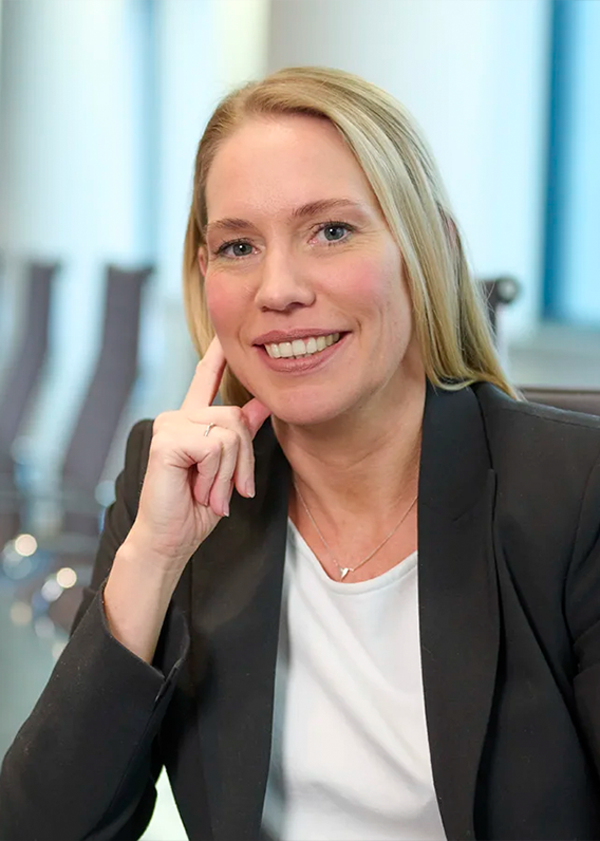 Eine lächelnde Frau in schwarzem Blazer und weißem Hemd sitzt an einem Tisch und stützt ihren Kopf auf ihre Hand.