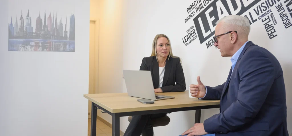 Zwei Personen in Business-Kleidung diskutieren Coaching Führungskräfte an einem Schreibtisch in einem modernen Büro mit motivierendem Wandtext.