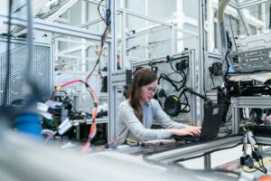 Frauen in der Produktion: Eine Frau arbeitet an einem Laptop, umgeben von Maschinen und Kabeln in einem modernen Industrielabor.