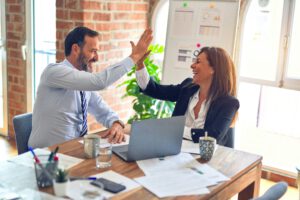 Zwei Kolleginnen, die an einem Schreibtisch lächeln und sich abklatschen – perfekte Teamarbeit in „Coaching für Frauen in Führungspositionen“.