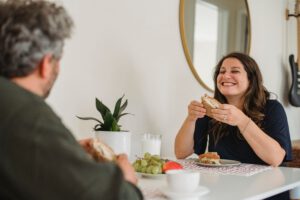 Frauen in der „Sandwich-Position“: Eine Frau lächelt, hält ein Sandwich in der Hand und genießt an einem Tisch das Mittagessen in Gesellschaft.