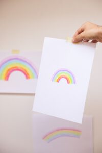 Eine Hand hält eine Regenbogenzeichnung hoch, die kreative Einblicke in MDI-Erfahrungen widerspiegelt; im Hintergrund sind weitere Regenbogen zu sehen.