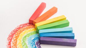 Sechs bunte Kreidestücke, sortiert nach Farben, mit denen man einen Regenbogenbogen auf weißes Papier malt.