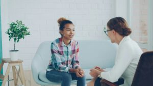 Zwei Frauen sitzen auf einem Sofa und unterhalten sich. Eine von ihnen hält ein Notizbuch und einen Stift in der Hand – möglicherweise befinden sie sich in einer Therapiesitzung.
